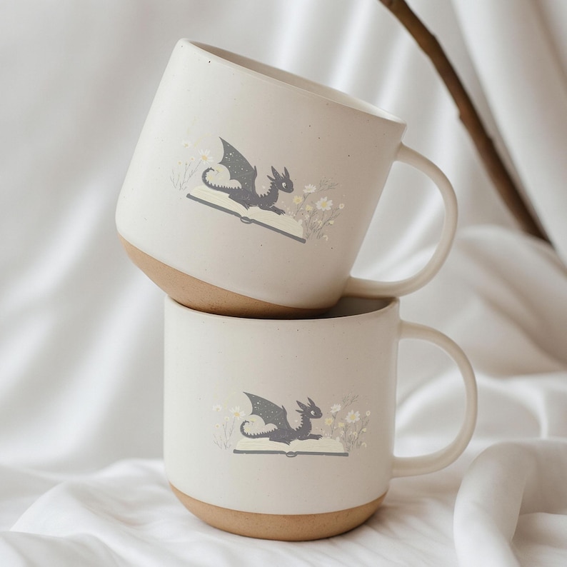 May include: Two off-white ceramic mugs with a dragon illustration. The dragon is gray and is depicted on an open book with flowers. The mugs have a brown base and a curved handle.