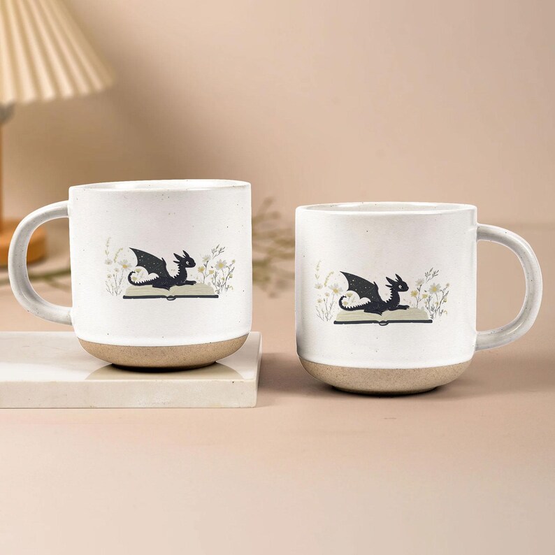 May include: Two white ceramic mugs with a dragon illustration. The dragon is black with outstretched wings, perched on an open book, surrounded by floral accents. The bottom of the mugs is a natural beige color.