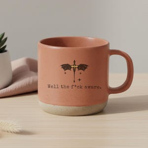 May include: A terracotta-colored ceramic mug with a dragon graphic and the text "Well the f*ck aware." The mug has a speckled finish and a cream-colored base. The handle is rounded and the mug is on a light-colored surface.