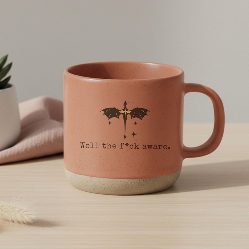 May include: A terracotta-colored ceramic mug with a dragon graphic and the text "Well the f*ck aware." The mug has a speckled finish and a cream-colored base. The handle is rounded and the mug is on a light-colored surface.
