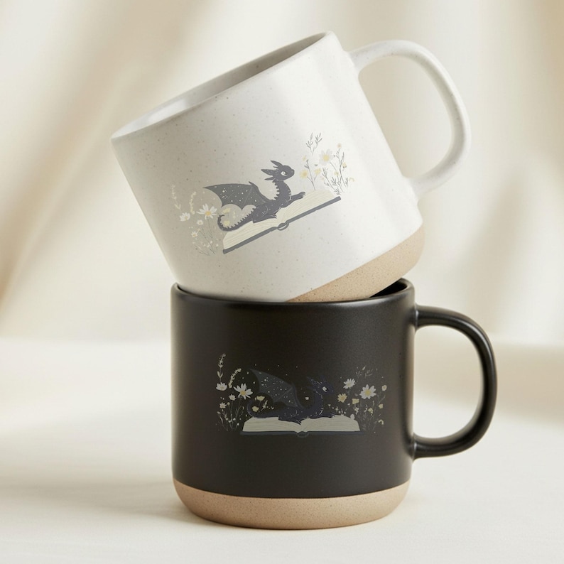 May include: Two ceramic mugs stacked on top of each other. The top mug is off-white with a dragon illustration on an open book. The bottom mug is black with a similar dragon design. Both mugs have a tan base and a curved handle.
