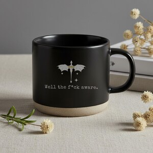 May include: Black ceramic mug with a beige base and a black handle. The mug features a white dragon graphic and the text "Well the f*ck aware."