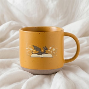 May include: Mustard yellow ceramic mug with a brown base and handle. The mug features a whimsical illustration of a black dragon resting on an open book, surrounded by white flowers. A charming addition to any kitchen.