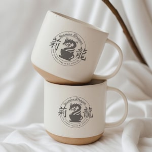 May include: Two white ceramic mugs stacked. Each mug has a tan base and a large handle. The mugs feature a circular logo with the text "Jasmine Dragon" and Chinese characters, along with the phrase "THE FINEST TEAS IN BA SING SE."