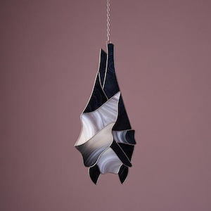 Può includere: Un ornamento a forma di pipistrello in vetro colorato appeso a una catena d'argento. Il pipistrello presenta un mix di pezzi di vetro nero, bianco e grigio, creando un effetto marmorizzato. Le ali e il corpo sono delineate con metallo argentato. L'opera è sospesa su uno sfondo viola.