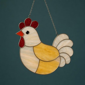 May include: Stained glass rooster suncatcher in white, yellow, and red. The rooster features a red comb and wattle, a yellow body, and white head and tail feathers. It is suspended by a metal chain.