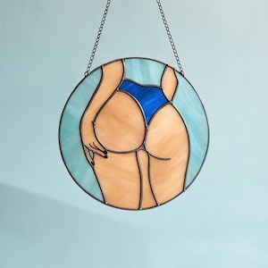 May include: Circular stained glass artwork showing a figure in a blue swimsuit. The piece has a light blue background and a chain for hanging. The figure's skin tones are a mix of peach and tan.