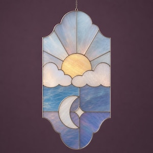 May include: Stained glass panel depicting a celestial scene. The design features a yellow sun with radiating rays, fluffy white clouds, and a blue sky. A crescent moon and star are also included. The panel is framed with a decorative border and a hanging chain.