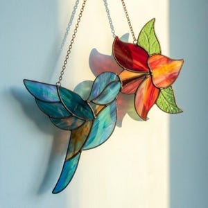 May include: Stained glass hummingbird and flower suncatcher. The hummingbird is in shades of blue, teal, and gold, while the flower features red, orange, and yellow petals with green leaves. Both are suspended by a chain.