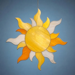 Op de afbeelding: Een gebrandschilderde zonnevanger met een heldergele kern en stralende vlammen in tinten geel, oranje en wit. Het zonmotief wordt opgehangen aan een ketting, perfect om een vleugje warmte en licht aan elke ruimte toe te voegen. Een decoratief item.