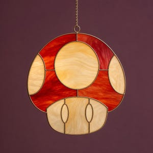 May include: A stained glass mushroom with a red and orange cap and a cream-colored stem. The mushroom is outlined in a gold-colored metal and is hanging from a chain. The mushroom is a decorative item.