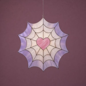 May include: A stained glass suncatcher shaped like a spiderweb, featuring a pink heart at the centre. The web design is white and lavender, framed in black metal. It hangs from a chain.