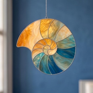 May include: A stained glass nautilus shell design in shades of orange, yellow, and blue, suspended by a silver chain. The spiral pattern is divided into sections, creating a colorful, decorative piece.