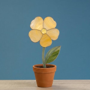 May include: A decorative yellow flower in a terracotta pot. The flower has five petals with a yellow center and a green stem and leaf. The pot is filled with soil and sits on a wooden surface against a blue background.