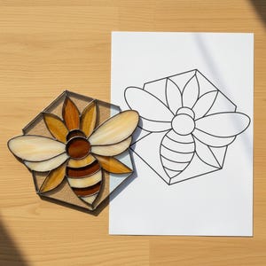 May include: A hexagonal stained glass bee suncatcher featuring amber, brown, and cream-colored glass segments joined by dark metal solder lines, displayed alongside a matching black and white line art pattern printed on a white sheet of paper.
