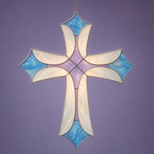 Pode incluir: Uma cruz de vitral com uma moldura de metal prateado. A cruz apresenta uma combinação de cores, incluindo azul claro, branco e lavanda. A cruz está pendurada por uma corrente sobre um fundo roxo.