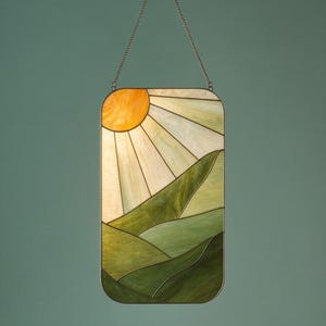 Könnte beinhalten: Buntglasfenster mit einer Landschaft mit gelber Sonne und grünen Hügeln. Das rechteckige Panel wird von einer Metallkette gehalten. Die Sonnenstrahlen sind hellgelb und die Hügel variieren in Grüntönen.