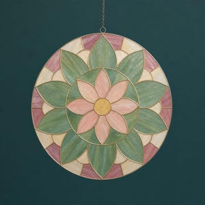 May include: A round, stained-glass style wall hanging with a floral design. The piece features a central flower with pink petals and a yellow center, surrounded by green leaves and pink and cream-colored segments, all outlined in gold.