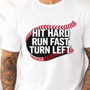 Op de afbeelding: Wit T-shirt met de tekst "HIT HARD RUN FAST TURN LEFT" in zwart en wit, ontworpen als een honkbal. Het shirt heeft ook een rood honkbalstikselontwerp. Het shirt wordt door een persoon gedragen.