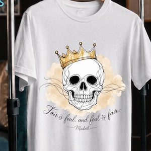 Puede incluir: Camiseta blanca con una calavera con una corona dorada y la cita "Fair is foul, and foul is fair" de Macbeth. El diseño incluye un fondo en forma de nube marrón claro y arte lineal negro.