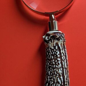 Può includere: Una lente d'ingrandimento con una lente circolare incorniciata in metallo dorato lucido, fissata a un manico realizzato con un pezzo di corno naturale testurizzato, marrone scuro e bianco, con una superficie dal motivo rugoso.