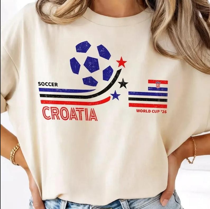 Pode incluir: Camiseta creme com um gr&aacute;fico de bola de futebol azul e uma estrela vermelha. O texto "SOCCER CROATIA" &eacute; impresso em vermelho e preto. Uma bandeira croata e o texto "WORLD CUP '26" tamb&eacute;m s&atilde;o vis&iacute;veis.