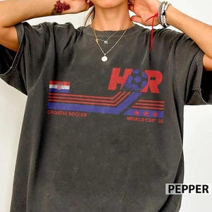 Peut inclure: Un t-shirt gris anthracite &agrave; manches courtes avec un imprim&eacute; graphique portant le texte HR, Croatia Soccer et World Cup 26. Le motif comprend un petit drapeau croate, des rayures horizontales rouges et bleues, un ballon et trois &eacute;toiles rouges.
