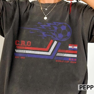 May include: Dark grey t-shirt with a soccer ball graphic and blue lightning. The shirt displays the text "C.R.O" in red, "EST. 1912" and "WORLD CUP 2026". Includes a Croatian flag design.