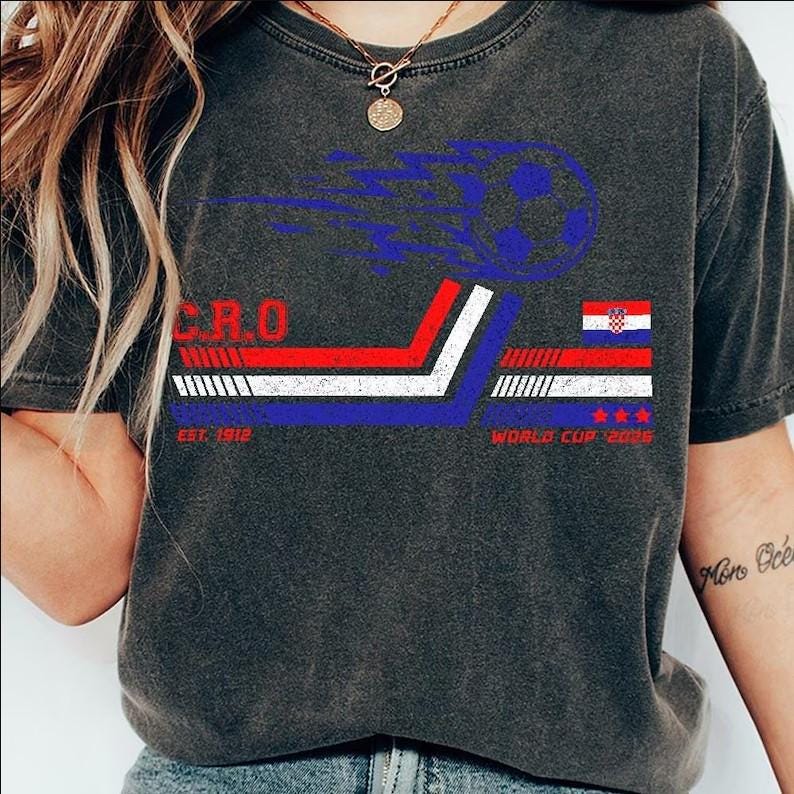 May include: Dark grey t-shirt with a graphic design in red, white, and blue, featuring the word "CRO", a soccer ball, the Croatian flag, and the text "EST. 1912" and "WORLD CUP 2018".