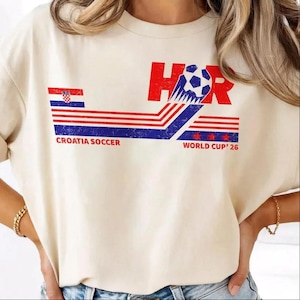 Peut inclure: Un t-shirt &agrave; manches courtes de couleur cr&egrave;me avec un motif graphique portant le texte Croatia Soccer et World Cup '26. Le design comprend un motif &agrave; rayures rouges, blanches et bleues, une ic&ocirc;ne de ballon de football et les lettres HR en rouge.