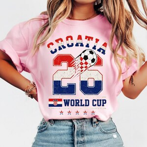 Może przedstawiać: Jasnor&oacute;żowy t-shirt z grafiką zawierającą napisy CROATIA oraz WORLD CUP. Wz&oacute;r przedstawia liczbę 26, piłkę nożną w szachownicę, małą flagę Chorwacji oraz pięć gwiazdek w kolorach czerwonym, białym i niebieskim.