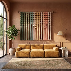 May include: A wall hanging featuring a horizontal wooden rod with multiple vertical strands of fabric spheres in a gradient of green, yellow, orange, and pink, each ending in a long tassel, suspended above a mustard yellow three-section sofa.