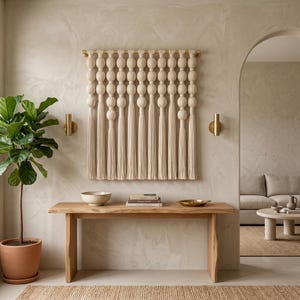 May include: A wall hanging featuring cream-colored fabric spheres suspended from a horizontal brass rod, with long, straight tassels extending downward from each vertical strand, creating a symmetrical and textured decorative textile piece.