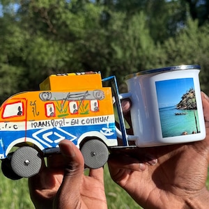 Peut inclure: Un bus jouet coloré et une tasse en émail blanche. Le bus est peint en jaune, bleu et orange avec l'inscription "TRANSPORT-EN COMMUN". La tasse présente une image d'une plage tropicale. Les objets sont tenus devant un fond vert.