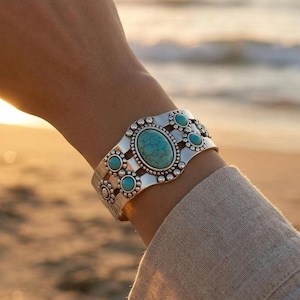 May include: A silver-toned cuff bracelet featuring a large central oval turquoise stone surrounded by smaller circular turquoise accents. The metal band displays a decorative beaded texture along the edges and intricate cut-out detailing.