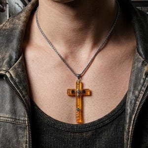 May include: A necklace featuring a translucent amber-colored cross pendant with metallic silver-toned accents and a central emblem, suspended from a silver-toned metal chain. The pendant has a rectangular cross shape with a smooth, polished finish.