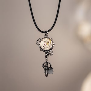 Può includere: Un ciondolo color argento a forma di chiave antica con un meccanismo di orologio circolare integrato che mostra ingranaggi dorati e argentati a vista, appeso a un semplice cordino nero che pende verticalmente su uno sfondo neutro.