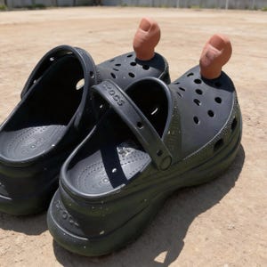 May include: Black Crocs clogs with a strap and a small, brown toe-shaped decoration in the front. The clogs are on a light brown surface. The word "Crocs" is visible on the side of the clogs.