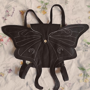 Può includere: Zaino nero a forma di falena con dettagli di cucitura bianchi. Lo zaino ha due ali curve, gambe a forma di antenna e una chiusura a bottone. Le ali presentano disegni a mezzaluna. Con due spallacci neri.