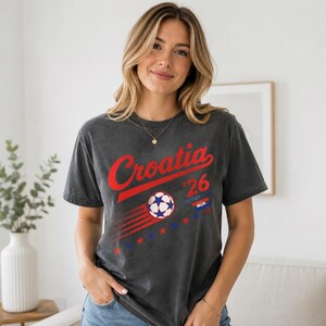 Puede incluir: Una camiseta de manga corta gris carb&oacute;n con el texto rojo Croatia, el n&uacute;mero 26, un gr&aacute;fico de bal&oacute;n de f&uacute;tbol con l&iacute;neas de movimiento, una peque&ntilde;a bandera croata y una serie de estrellas rojas y azules impresas en la parte delantera.