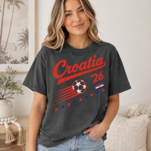 Puede incluir: Una camiseta de manga corta gris carb&oacute;n con el texto rojo Croatia, el n&uacute;mero 26, un gr&aacute;fico de bal&oacute;n de f&uacute;tbol con l&iacute;neas de movimiento, una peque&ntilde;a bandera croata y una fila de estrellas rojas y azules alternas en el pecho.