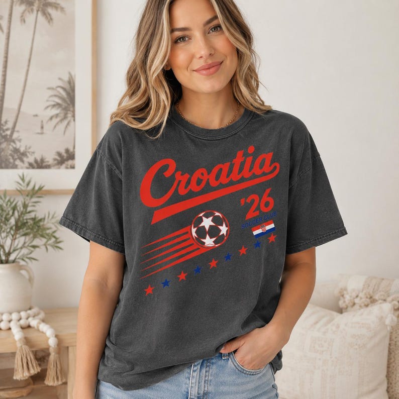Puede incluir: Una camiseta de manga corta gris carb&oacute;n con el texto rojo Croatia, el n&uacute;mero 26, un gr&aacute;fico de bal&oacute;n de f&uacute;tbol con l&iacute;neas de movimiento, una peque&ntilde;a bandera croata y una fila de estrellas rojas y azules alternas en el pecho.