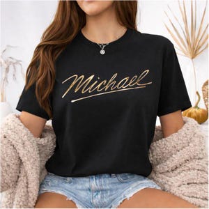 Op de afbeelding: Zwart T-shirt met de naam "Michael" in gouden letters. Het T-shirt heeft een ronde hals en is gemaakt van een zacht materiaal. De tekst is in een cursief lettertype.