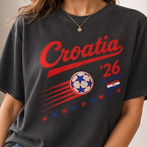 Puede incluir: Una camiseta gris carb&oacute;n con el texto rojo Croatia sobre un gr&aacute;fico de un bal&oacute;n de f&uacute;tbol con l&iacute;neas de movimiento. El dise&ntilde;o incluye el texto '26, las palabras SOCCER CLUB, una peque&ntilde;a bandera croata y una fila de estrellas rojas y azules.