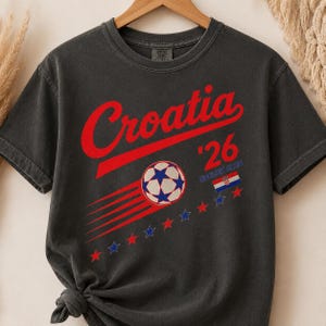 Puede incluir: Una camiseta de manga corta gris carb&oacute;n con el texto rojo Croatia, el n&uacute;mero 26 y las palabras SOCCER CLUB. El dise&ntilde;o incluye un bal&oacute;n de f&uacute;tbol con estrellas azules, l&iacute;neas de movimiento rojas, una peque&ntilde;a bandera croata y estrellas.