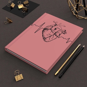 May include: A pink journal with a black illustration of a human heart and an EKG line. The journal is closed and has a hardcover. Gold and black pencils and gold binder clips are also visible.