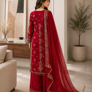 May include: A deep red long-sleeved tunic and wide-leg trousers set featuring intricate gold embroidery, sequin embellishments, and a matching sheer red dupatta with a decorative gold border. The garment displays detailed floral patterns throughout.