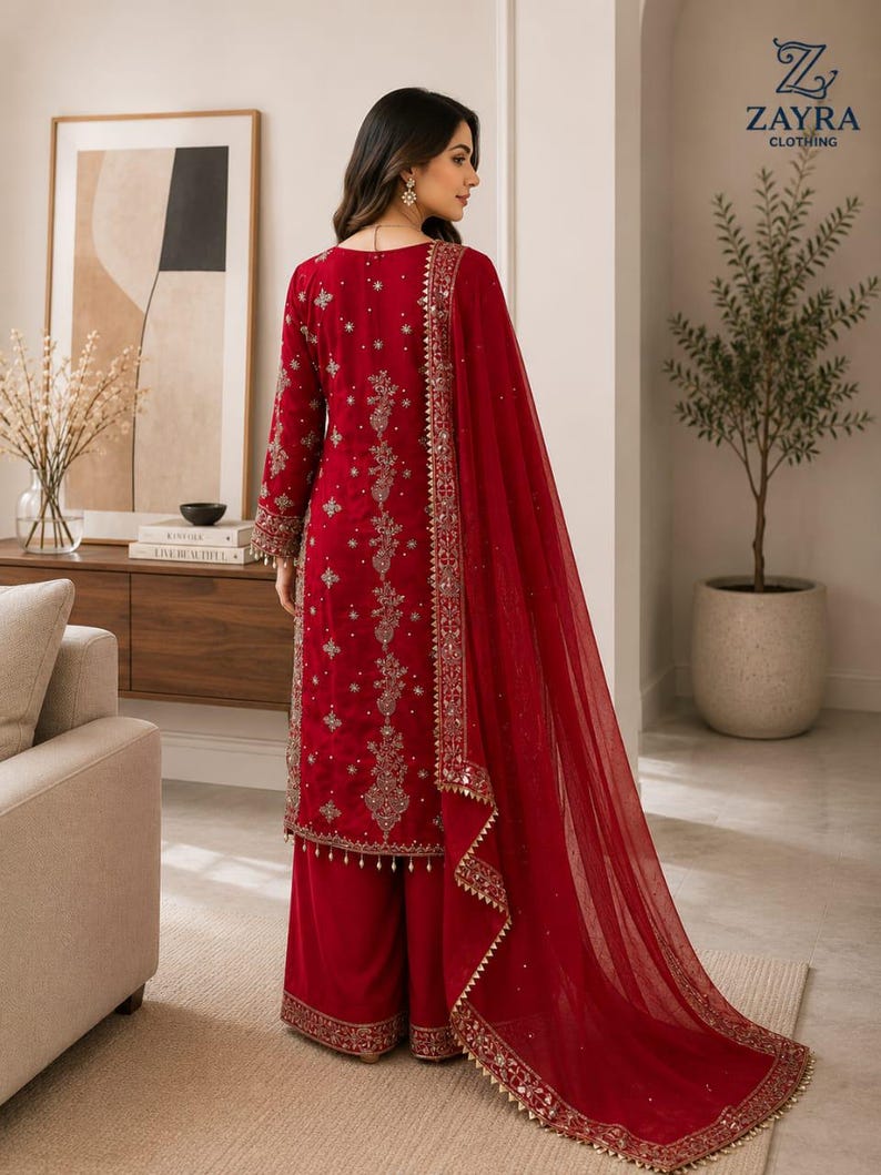 May include: A deep red long-sleeved tunic and wide-leg trousers set featuring intricate gold embroidery, sequin embellishments, and a matching sheer red dupatta with a decorative gold border. The garment displays detailed floral patterns throughout.