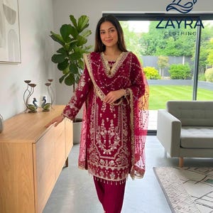 May include: A deep magenta long-sleeved tunic and matching trousers set featuring intricate white floral embroidery and gold-toned trim. The outfit includes a sheer, matching magenta dupatta with embroidered edges and a ZAYRA CLOTHING logo.
