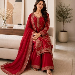 May include: A deep red long-sleeved tunic and wide-leg trousers set featuring intricate gold embroidery along the neckline, cuffs, and hem. The outfit includes a matching sheer red dupatta with a decorative gold border and small hanging beads.